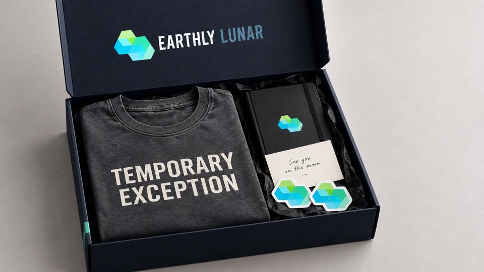 An open navy box with the Earthly Lunar logo inside the lid, holding a folded charcoal 'Temporary Exception' tee, a black notebook with a 'See you on the moon' card, and two hexagon stickers