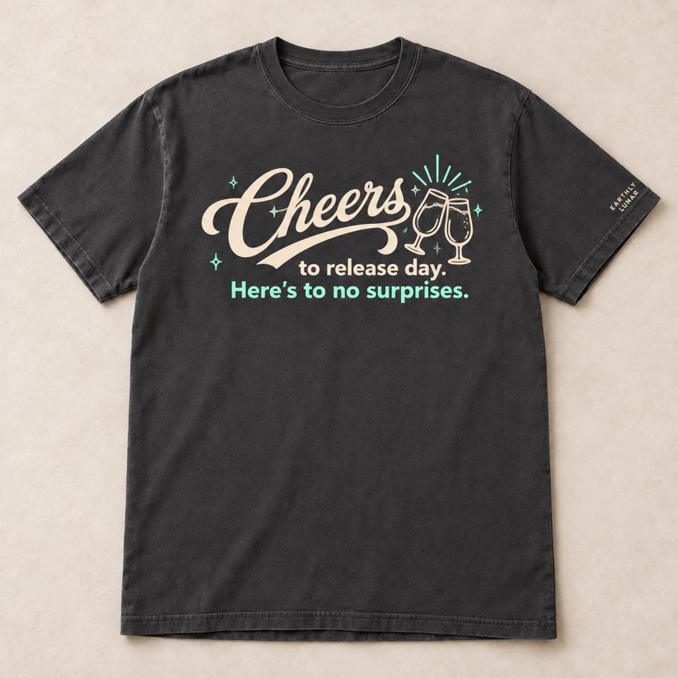 Charcoal tee with 'Cheers' in cream script beside two clinking glasses, over 'to release day.' and 'Here's to no surprises.' in mint